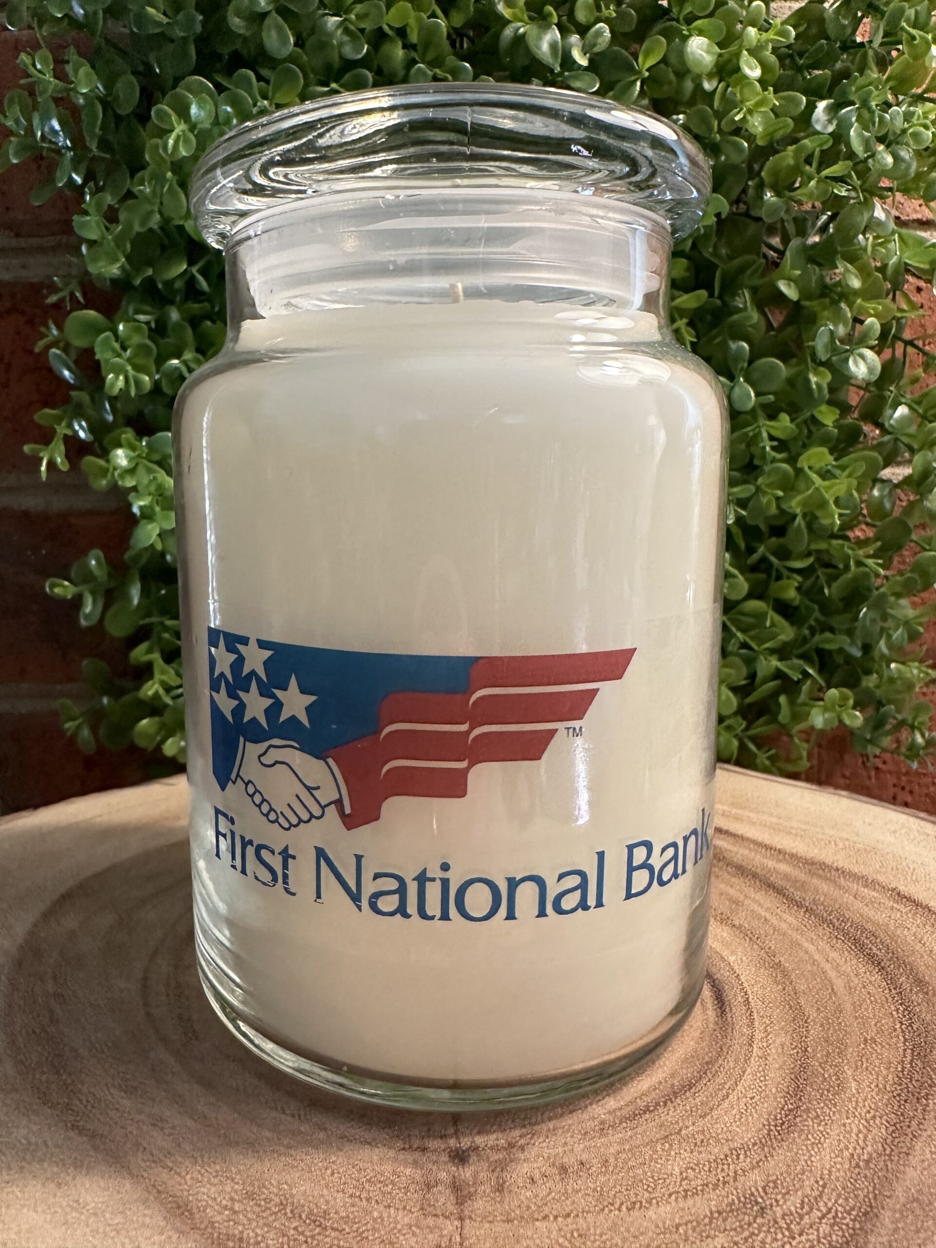 One of The Lighthouse Candle Factory's Custom Corporate Candle Gifts inside a clear glass jar with white wax featuring the First National Bank logo.